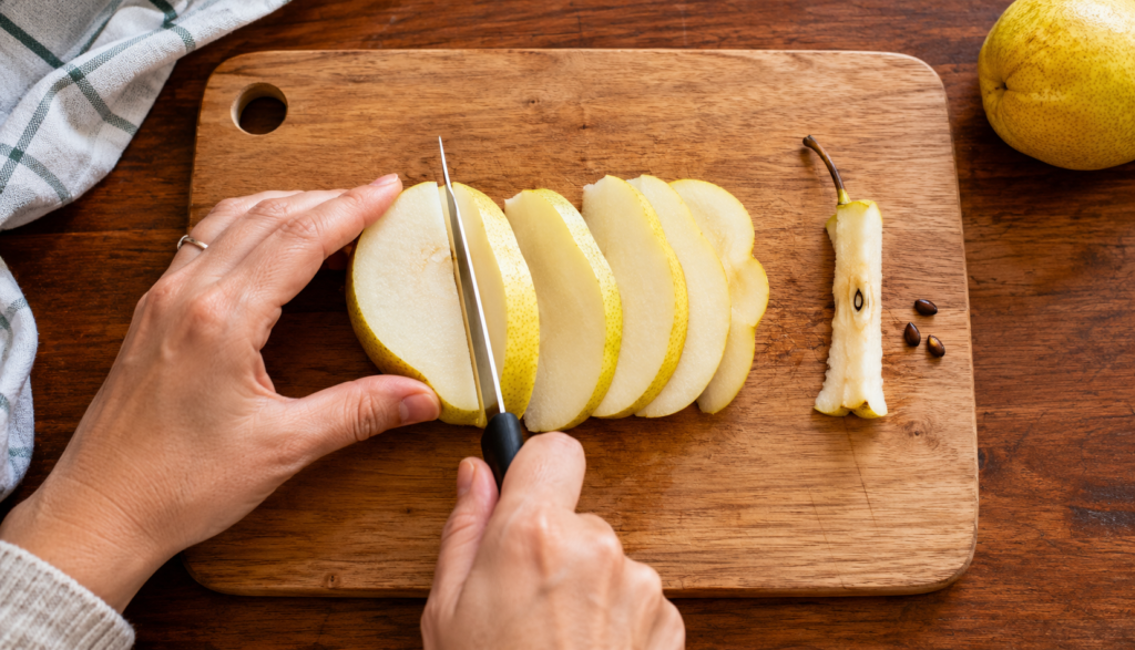 Hands slicing pear on cutting board with core and seeds removed