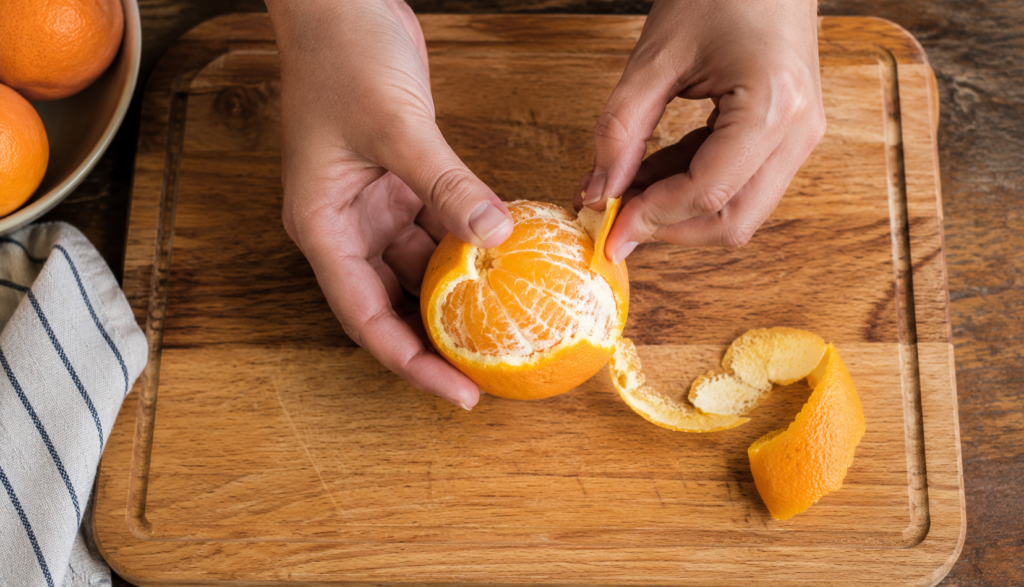 Hands peeling fresh orange over wooden cutting board showing flesh beneath